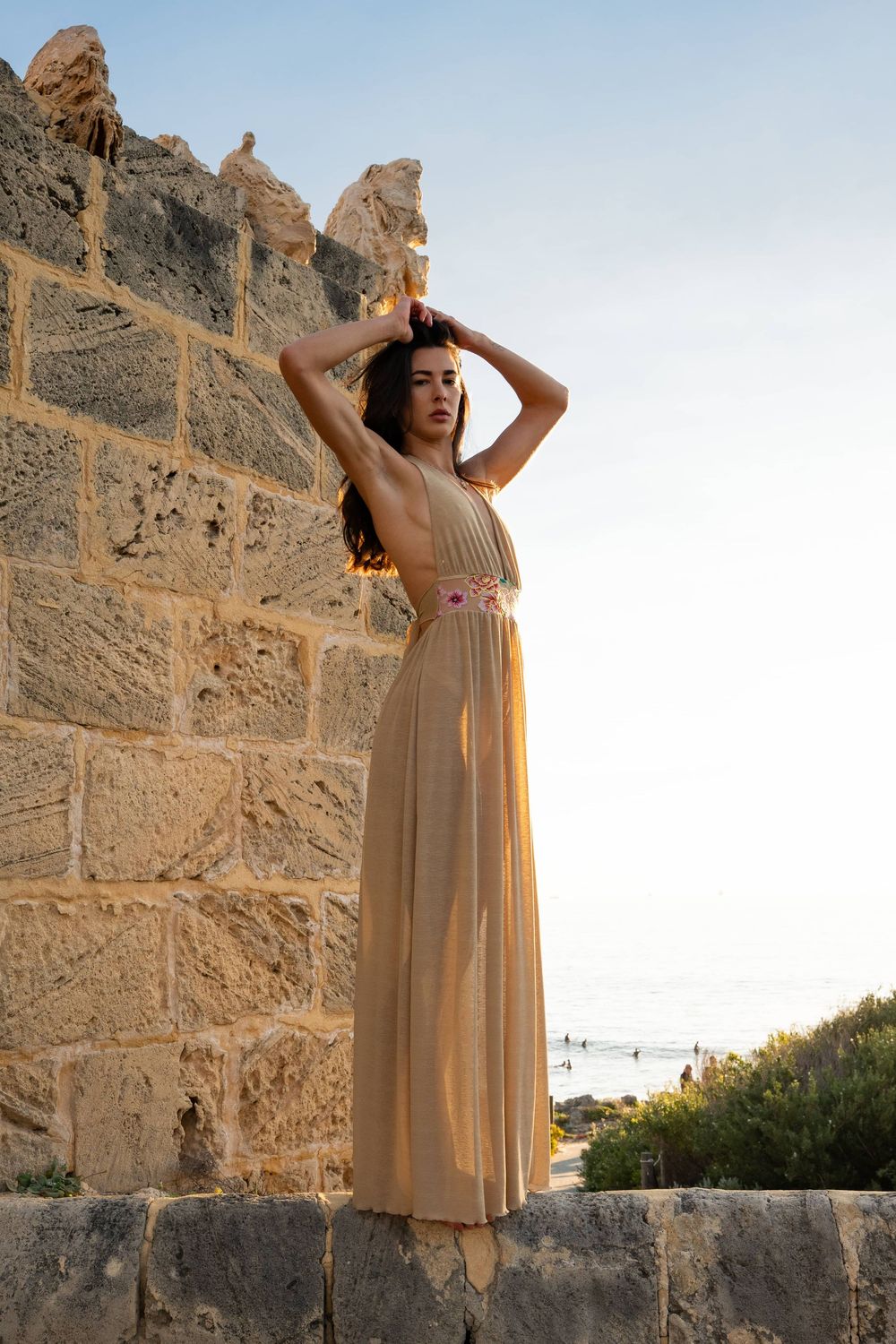 Woman in elegant beige maxi dress poses against ancient stone wall at sunset near the sea.