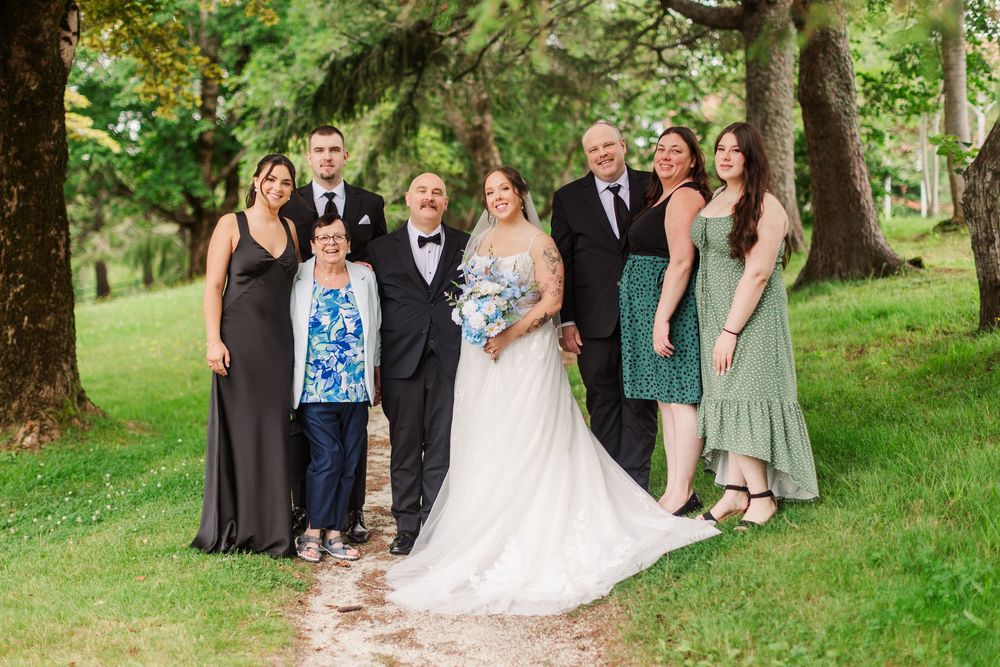 Wedding party poses outdoors under large trees on a gravel path, bride in white gown holding bouquet with family.