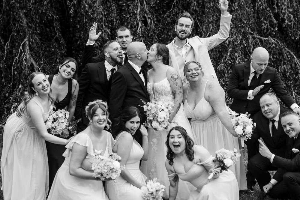 Black and white wedding party photo with bridesmaids holding bouquets and groomsmen celebrating joyfully outdoors.