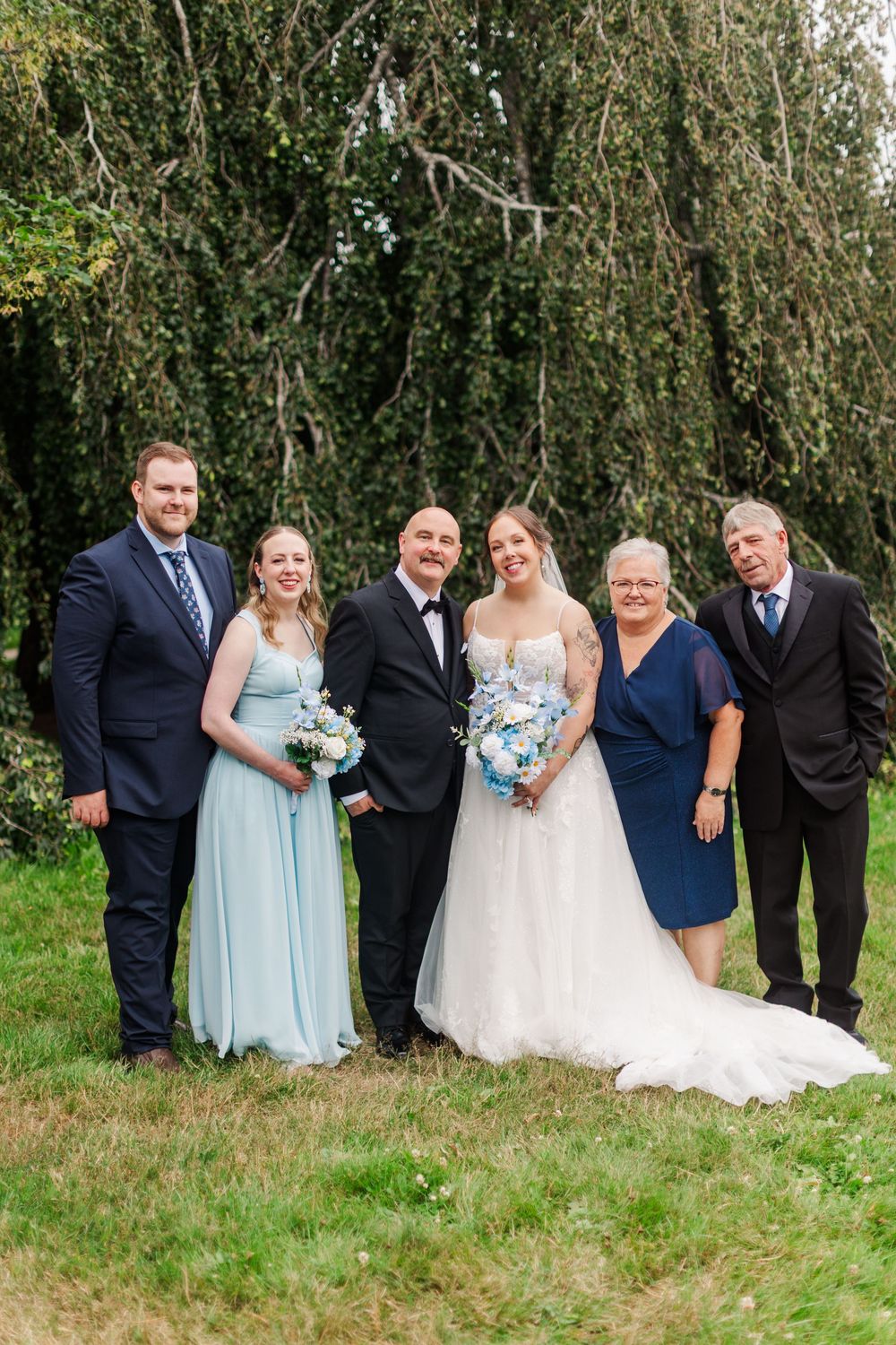 Wedding party poses outdoors with bride in white gown, groom in tuxedo, bridesmaid in blue, and family members.