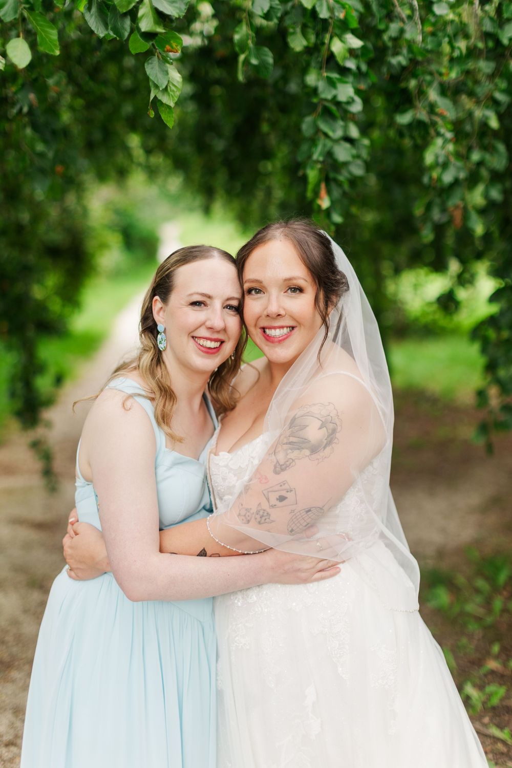 Bride in white gown and veil embraces bridesmaid in light blue dress under green tree canopy.