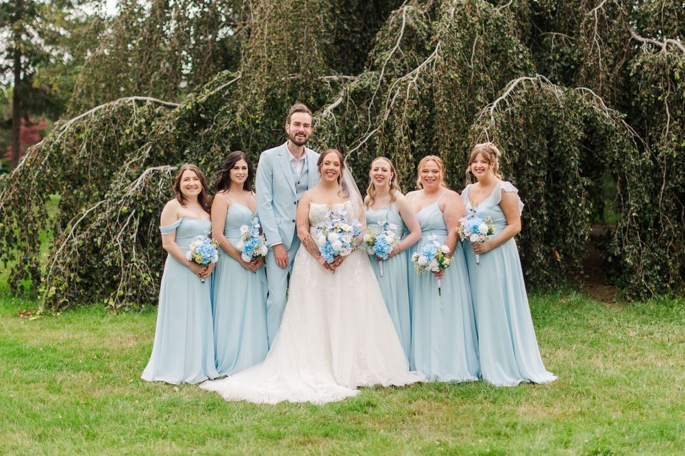 Bride and groom pose with bridesmaids in light blue gowns under a weeping tree at an outdoor wedding.