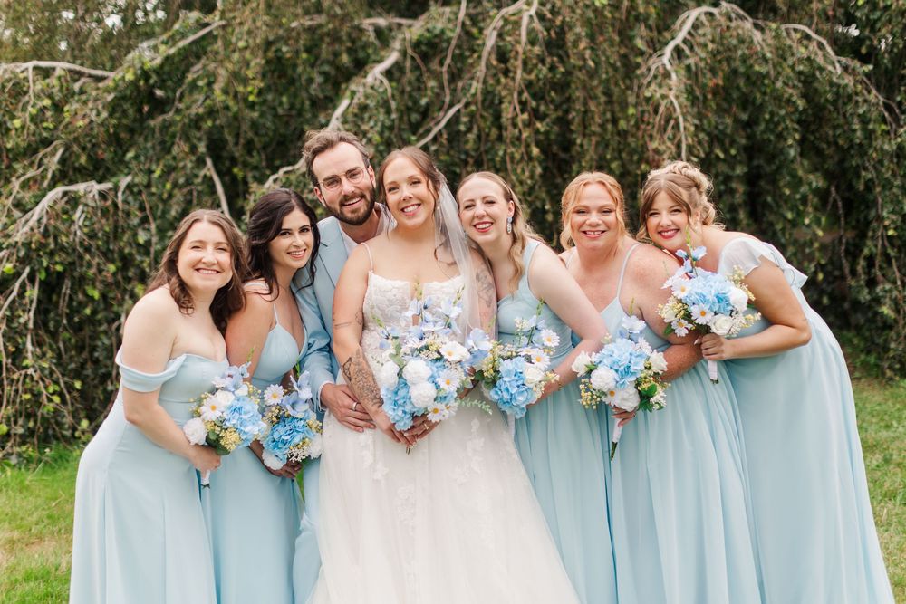 Bride and groom smile with five bridesmaids in light blue gowns holding blue floral bouquets outdoors.