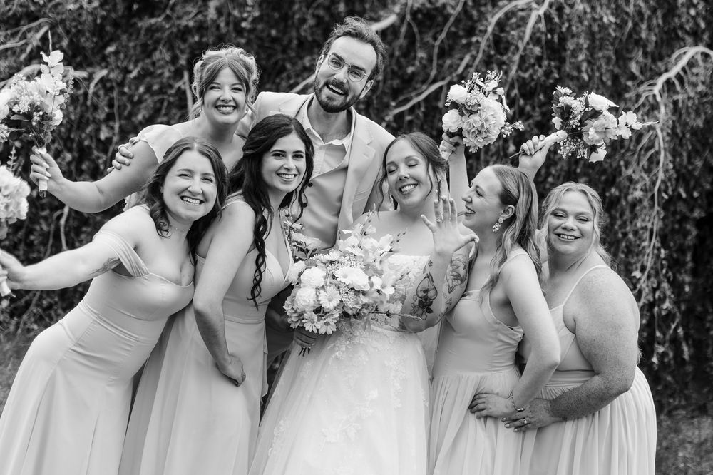 Black and white wedding photo of smiling bride with bouquet surrounded by five bridesmaids in light dresses and tall groom outdoors.