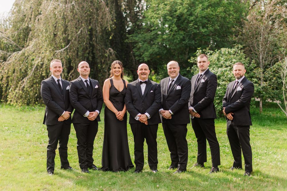 Wedding party in black formal attire posing outdoors on green grass with weeping willow trees in background.