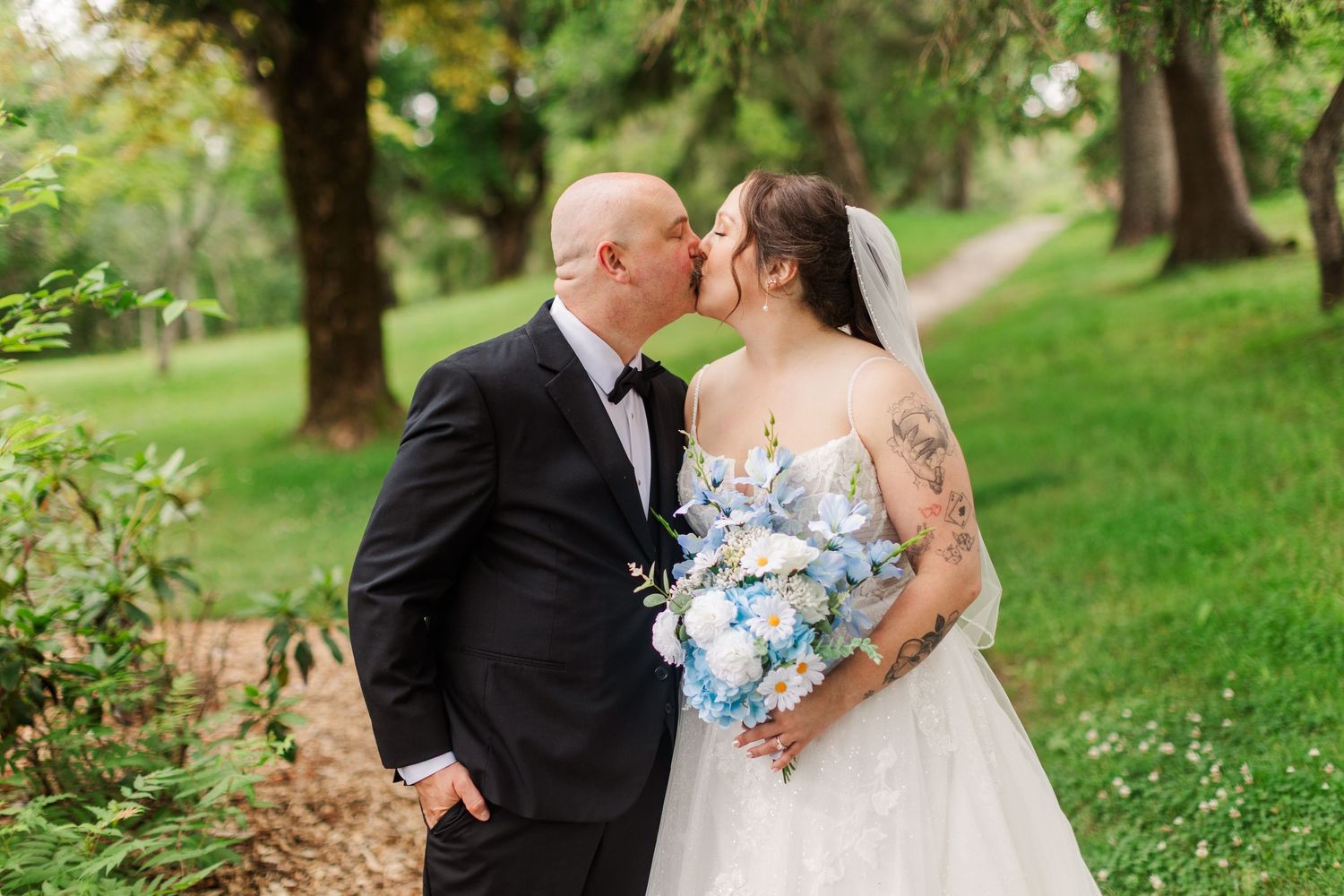 A bride and groom share a romantic kiss outdoors, surrounded by lush green trees, as she holds a blue and white bouquet.