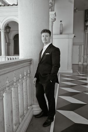 Man in black suit with pocket square poses confidently in elegant columned venue with geometric floor.
