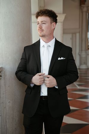 Young man in black suit with white tie and pocket square adjusts jacket buttons in elegant marble hallway.
