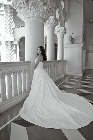 Bride in elegant white wedding gown with long train poses on ornate balcony with classical columns in black and white.