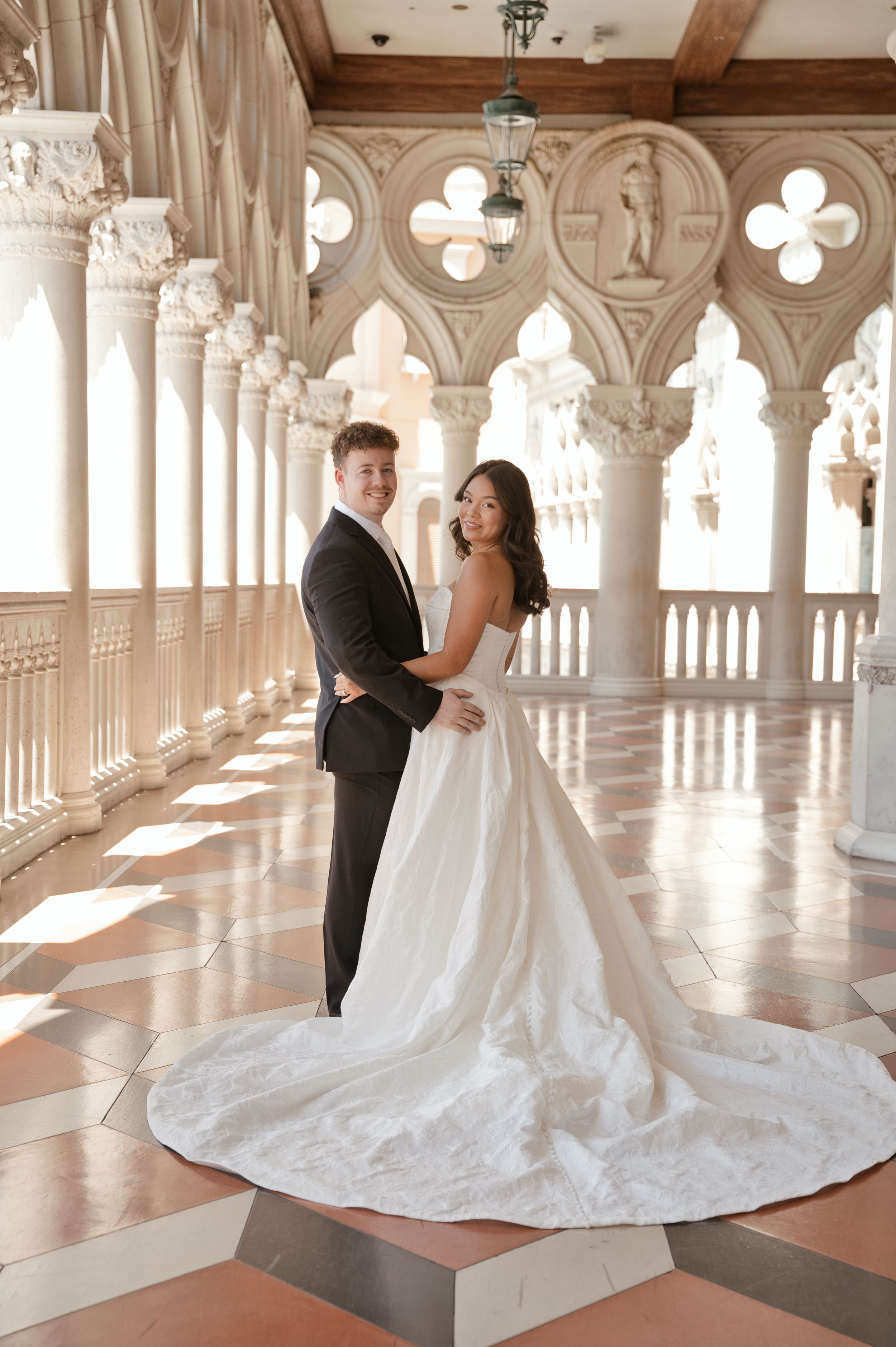 Bride and groom pose elegantly in a grand marble colonnade, her white ballgown train flowing across the ornate tiled floor.