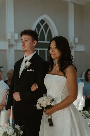 A bride and groom stand at the altar during their wedding ceremony, she holds white roses in a strapless gown.