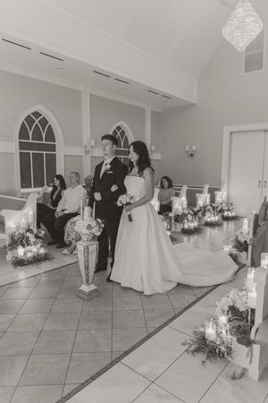 Black and white wedding photo of bride and groom walking down chapel aisle lined with candles and floral arrangements.