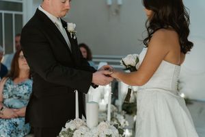 Groom places wedding ring on brides finger during intimate chapel ceremony with white floral decor.