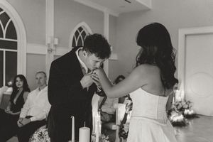 Groom kissing bride's hand at wedding altar in black and white photo with candles and guests in background.