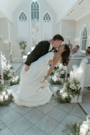 Bride and groom share a romantic dip kiss at the altar of a candlelit white chapel during their wedding ceremony.