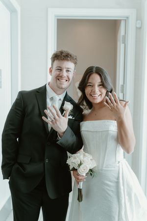 Newlywed couple smiling and showing off wedding rings, bride in white gown holding bouquet, groom in black suit.