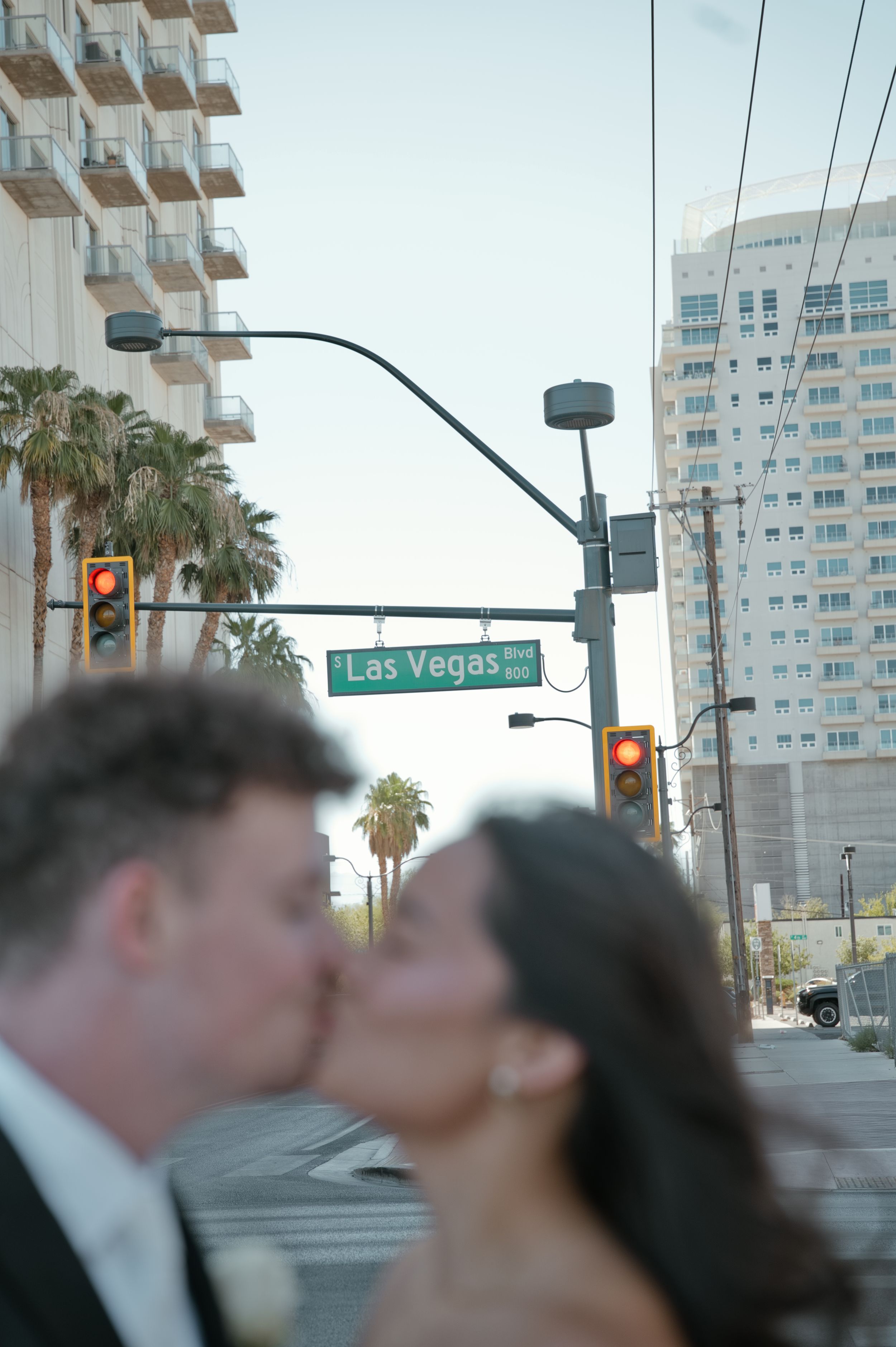 Newlyweds share a kiss at the Las Vegas Blvd street sign intersection with city buildings and palm trees in the background.