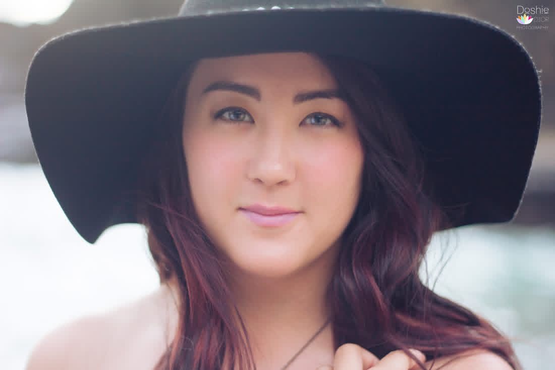 Young woman with long dark hair wearing a wide-brim black hat, posing outdoors in a soft-focus portrait.