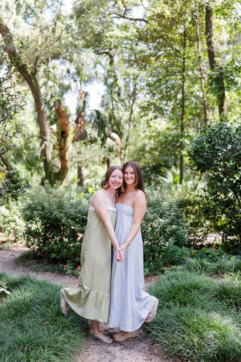 Two young women in flowy maxi dresses pose together smiling in a lush green garden path.