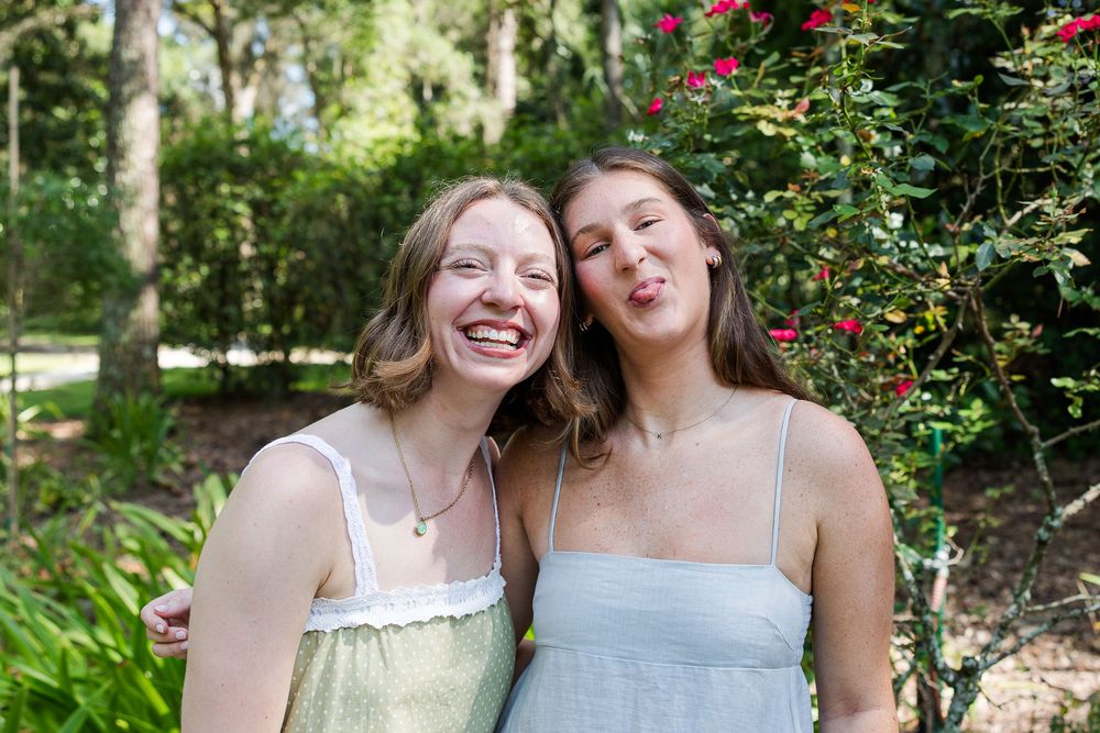 Two young women smiling and posing together outdoors in a lush green garden with red flowers.