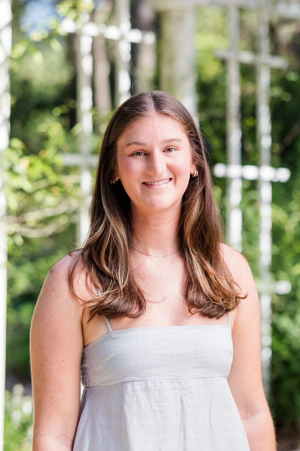 Smiling young woman with long brown hair wearing a light gray sundress outdoors in a garden setting.