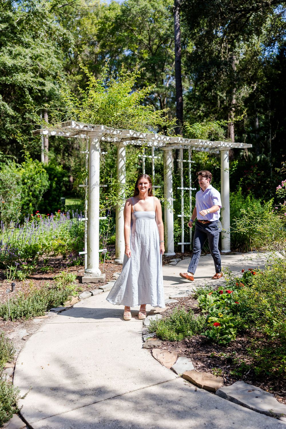 Woman in white sundress walks garden path near white pergola with man following behind.