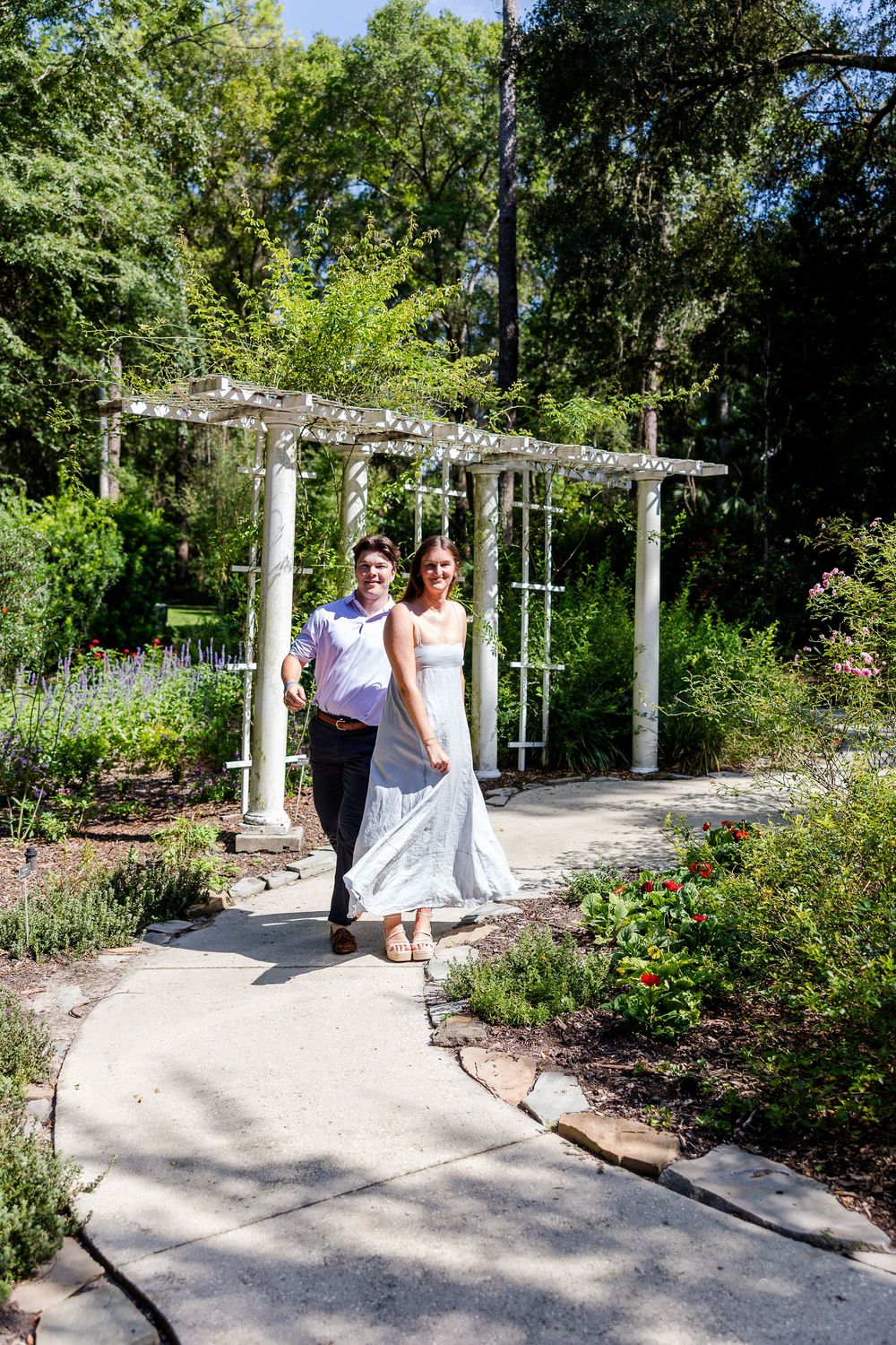 Couple walks hand-in-hand along a garden path under a white pergola surrounded by lush greenery.
