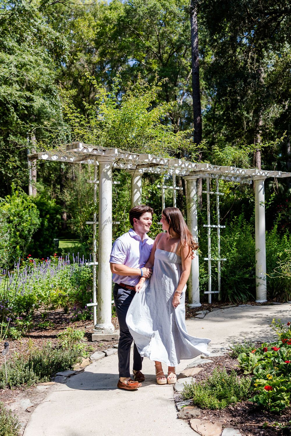 Couple shares romantic moment on garden path beside white pergola surrounded by lush greenery.