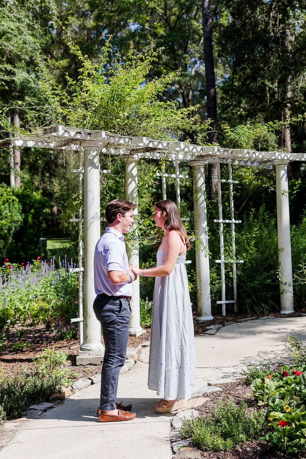 Couple holding hands facing each other under a white garden pergola surrounded by lush greenery and colorful flowers.