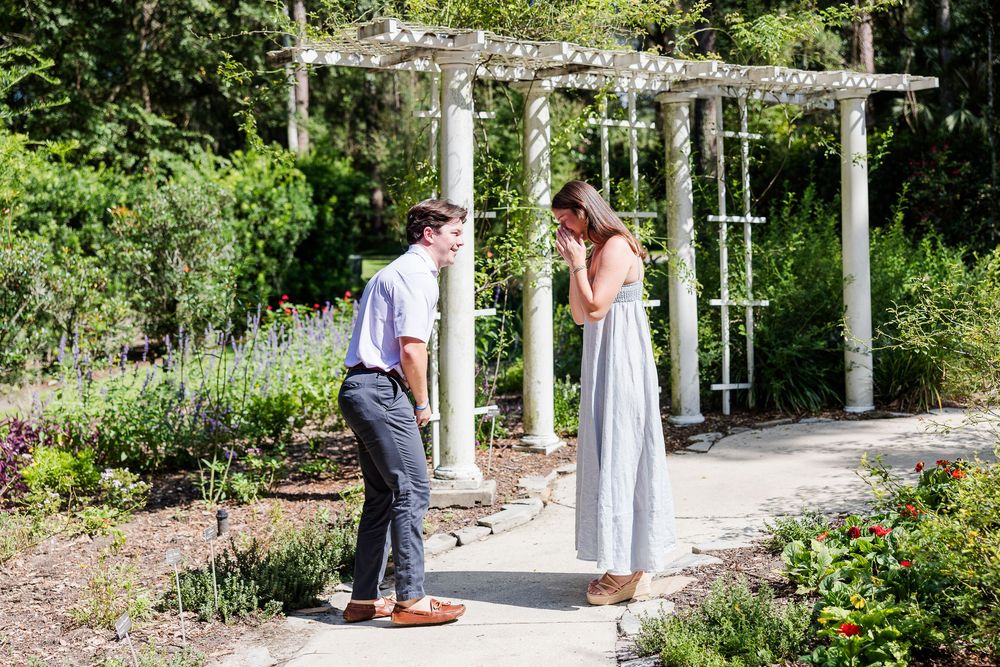 Couple sharing an emotional moment on a garden path under a white pergola surrounded by colorful flowers and greenery.