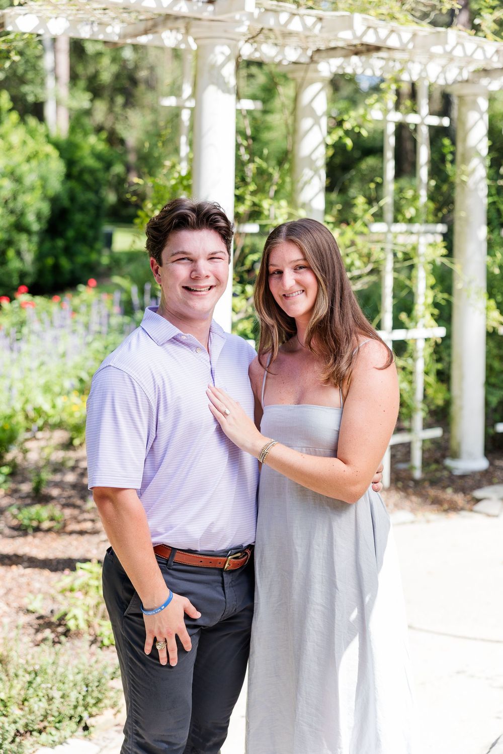 Smiling couple posing together in a lush garden with a white pergola backdrop on a sunny day.