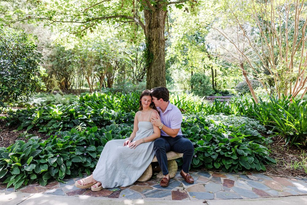 Couple sitting on a stone bench surrounded by lush greenery in a romantic garden engagement photo.
