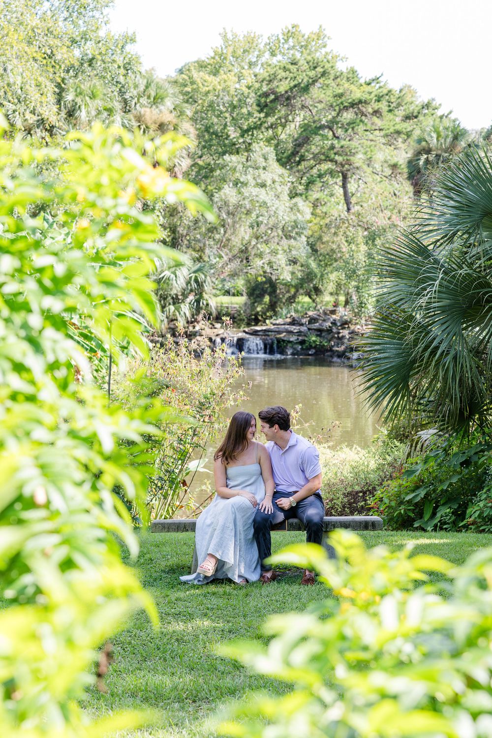 Couple sitting close together on a bench by a serene pond surrounded by lush greenery and tropical plants.