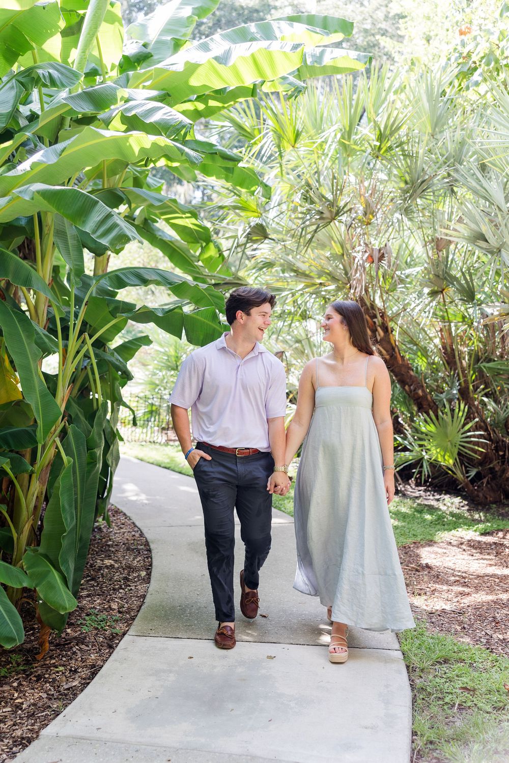 Young couple walks hand-in-hand on a path lined with tropical banana plants and palm trees.