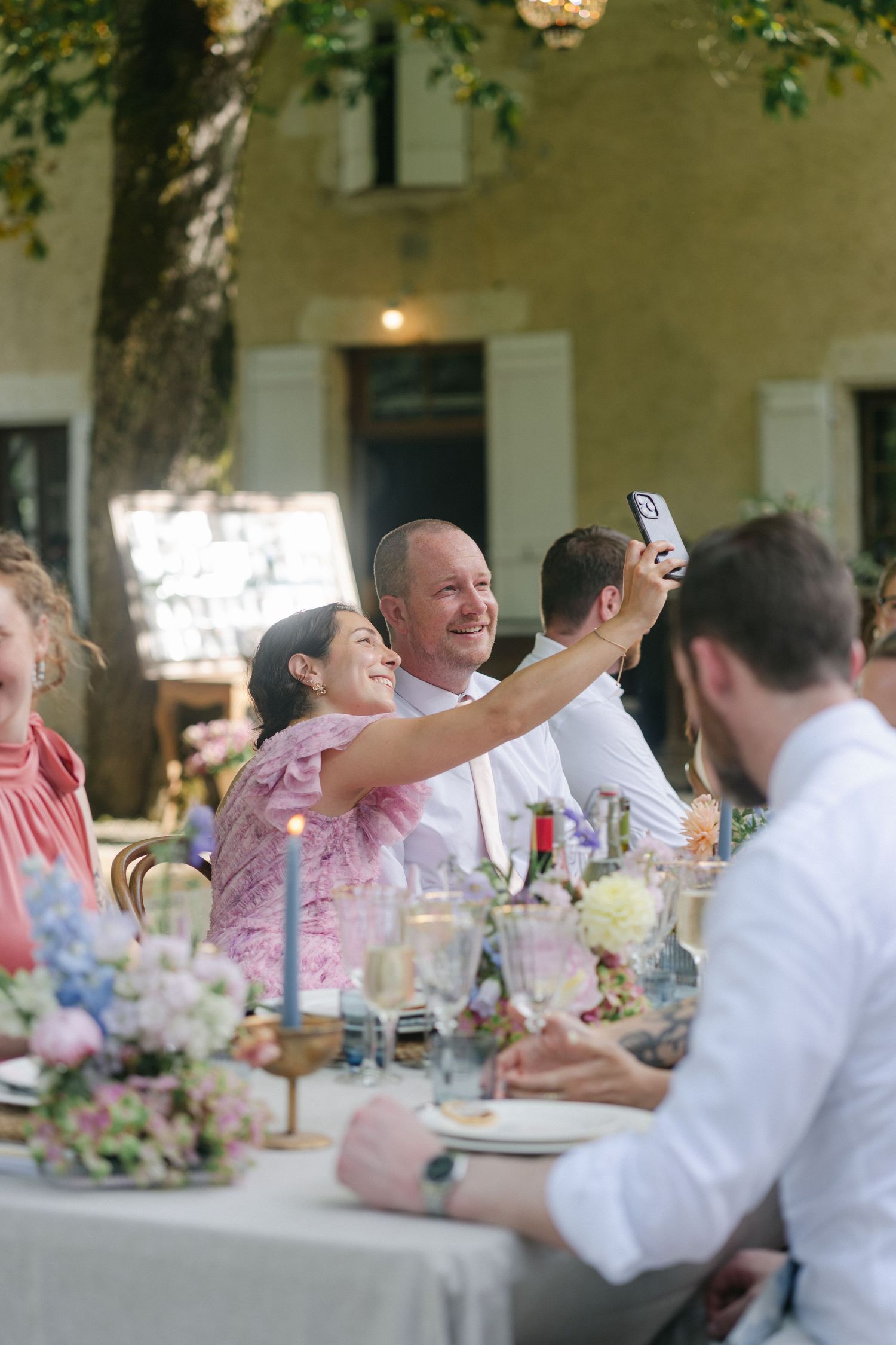 Guests taking a selfie at an elegant outdoor wedding reception with floral centerpieces and candlelit tables.