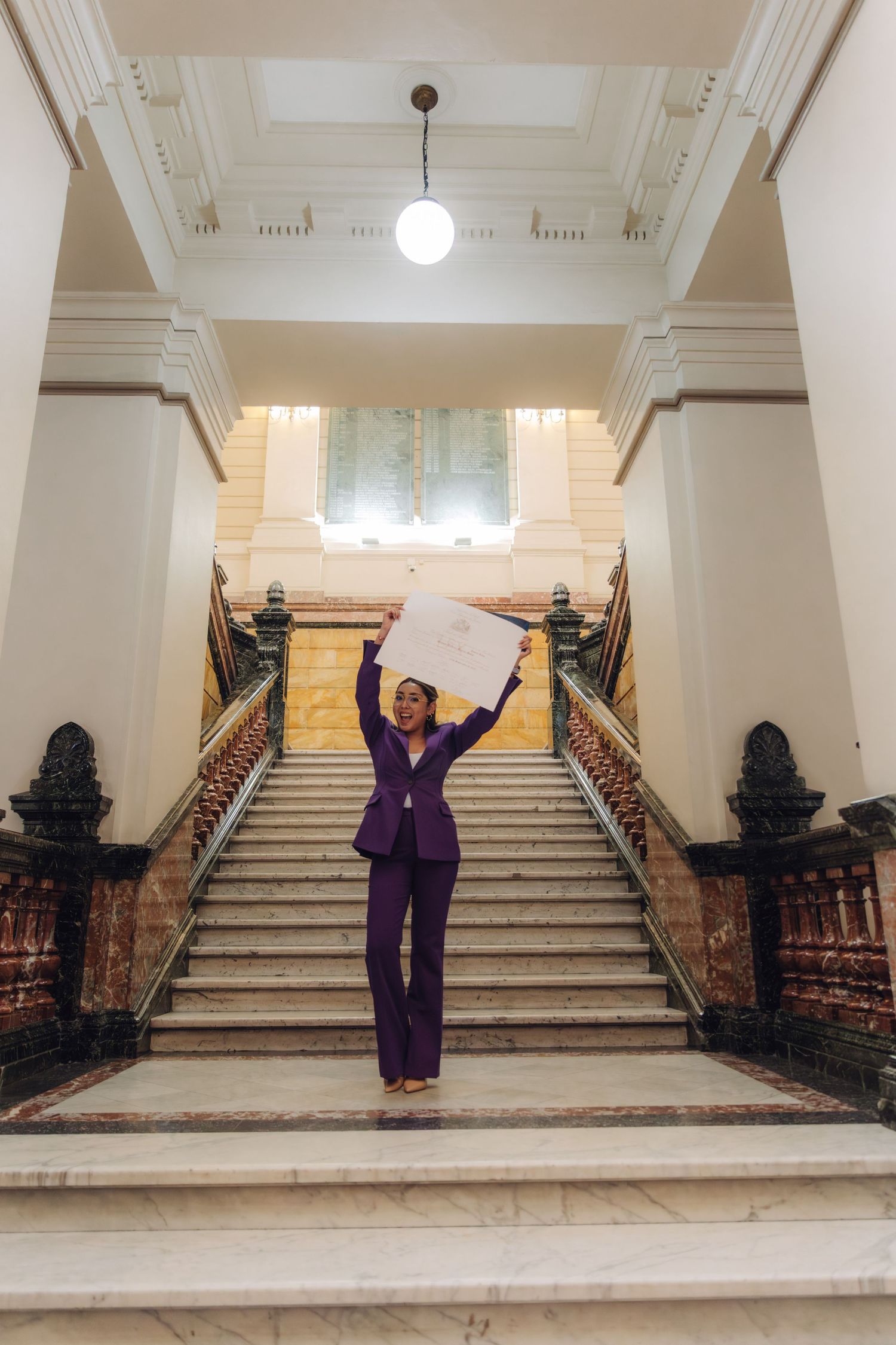 Woman in purple suit triumphantly raising certificate on grand marble staircase.
