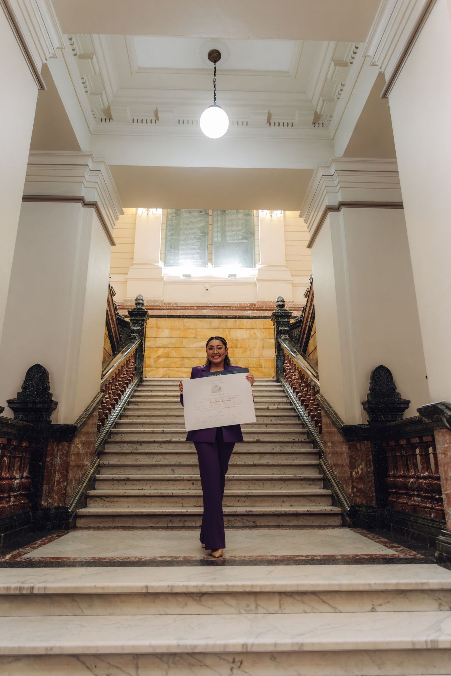 Woman in purple suit descending ornate marble staircase holding official certificate.