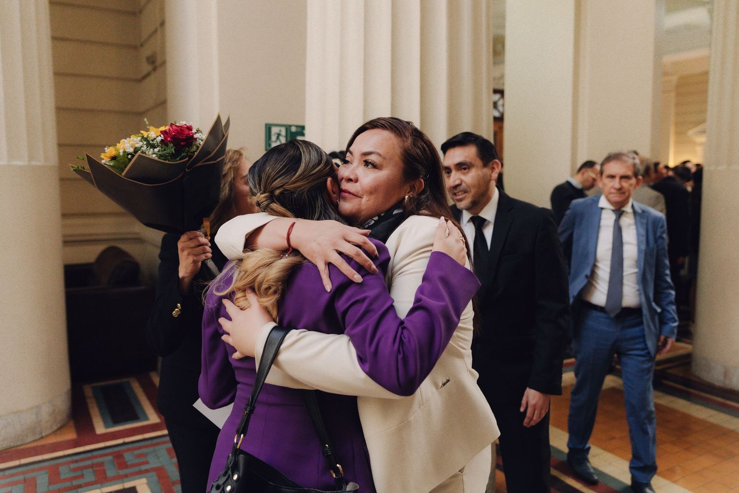 Two women sharing a warm embrace in elegant columned hallway with flowers.