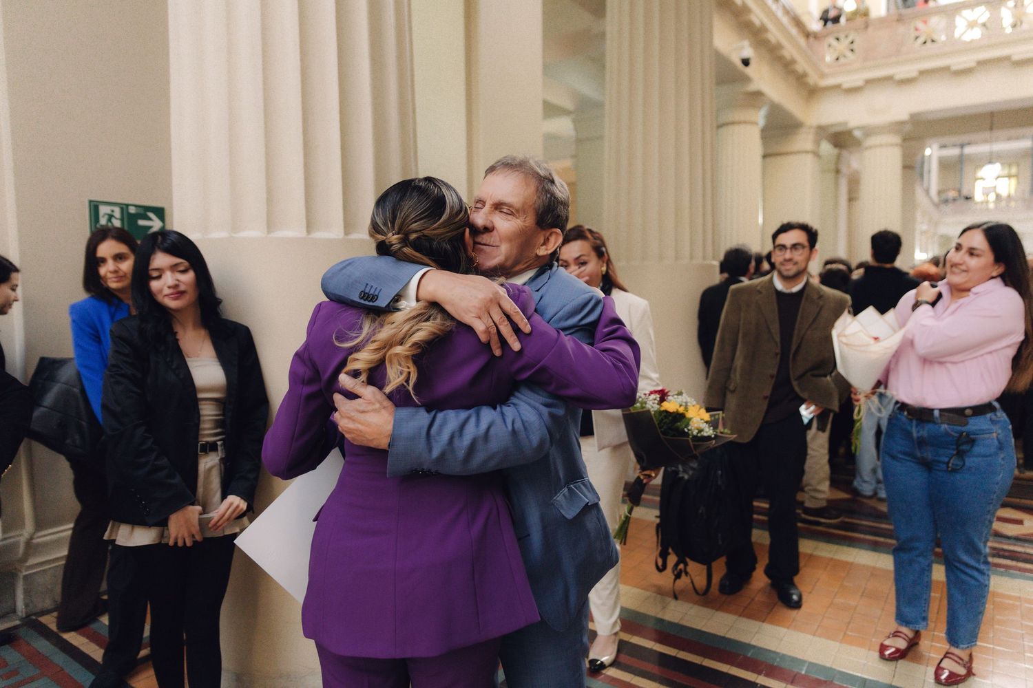 Woman in purple suit hugging man in elegant columned government building hallway.