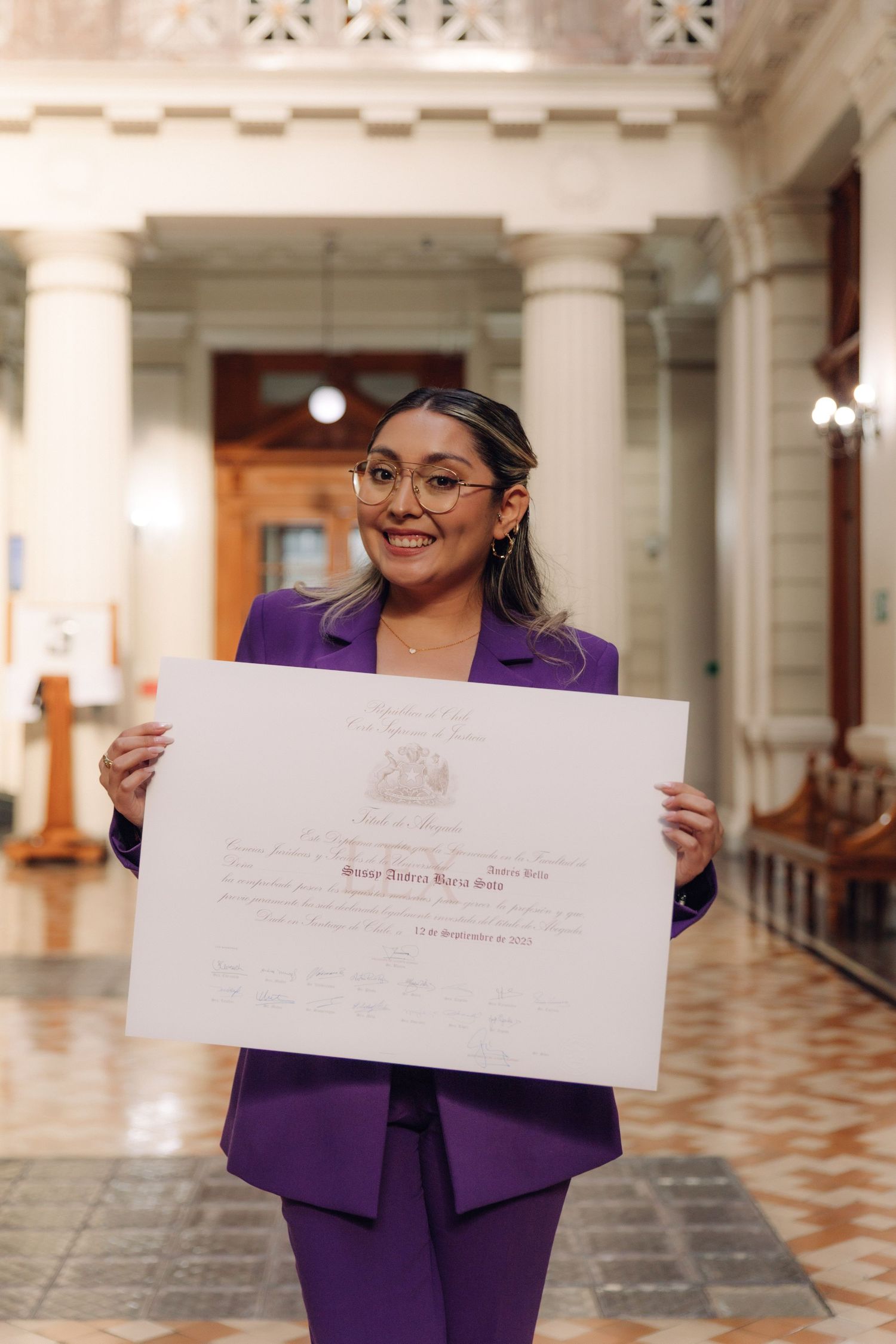 Smiling woman in purple suit displaying official certificate in grand columned hall.