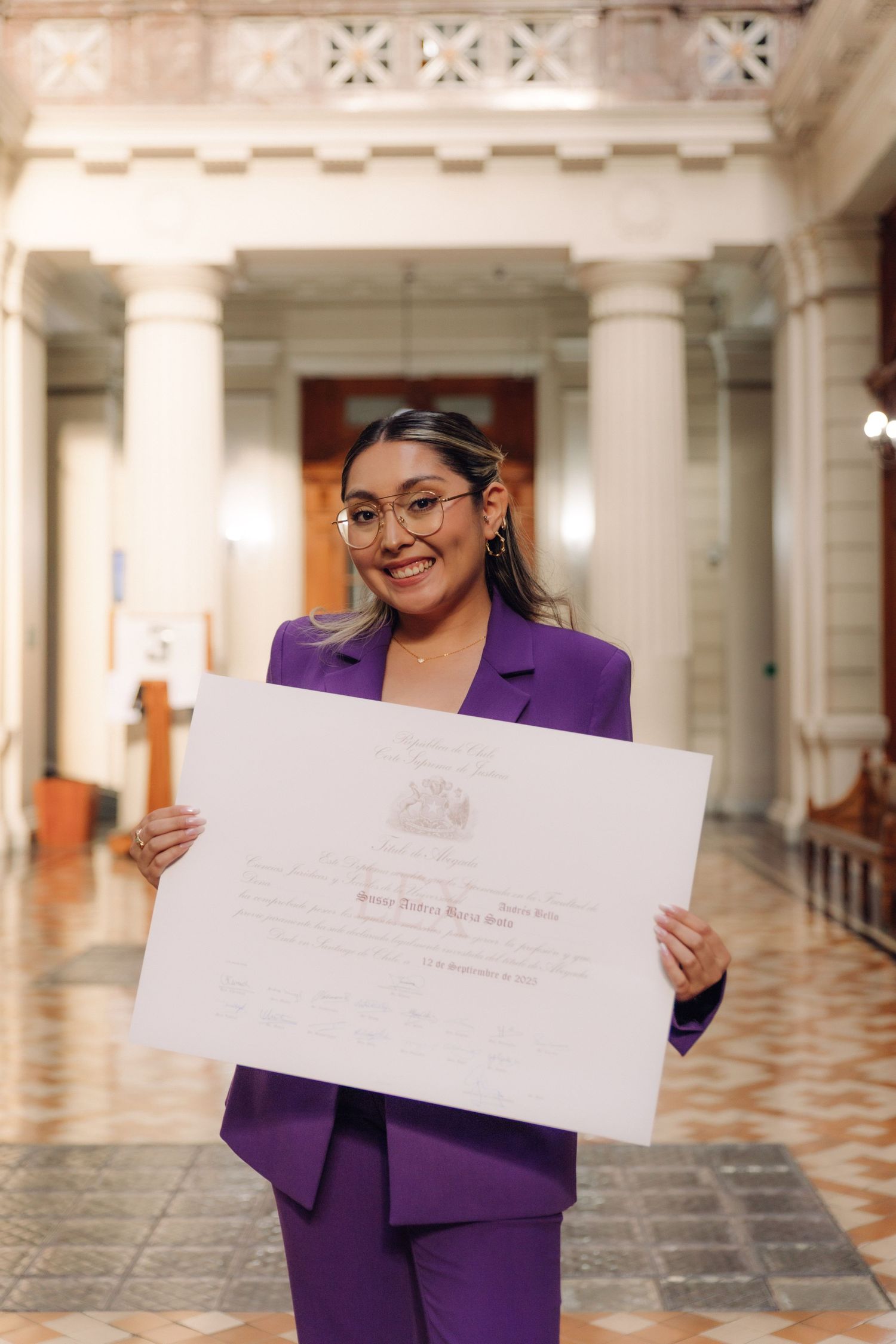 Smiling woman in purple suit holding large official certificate in grand hall.