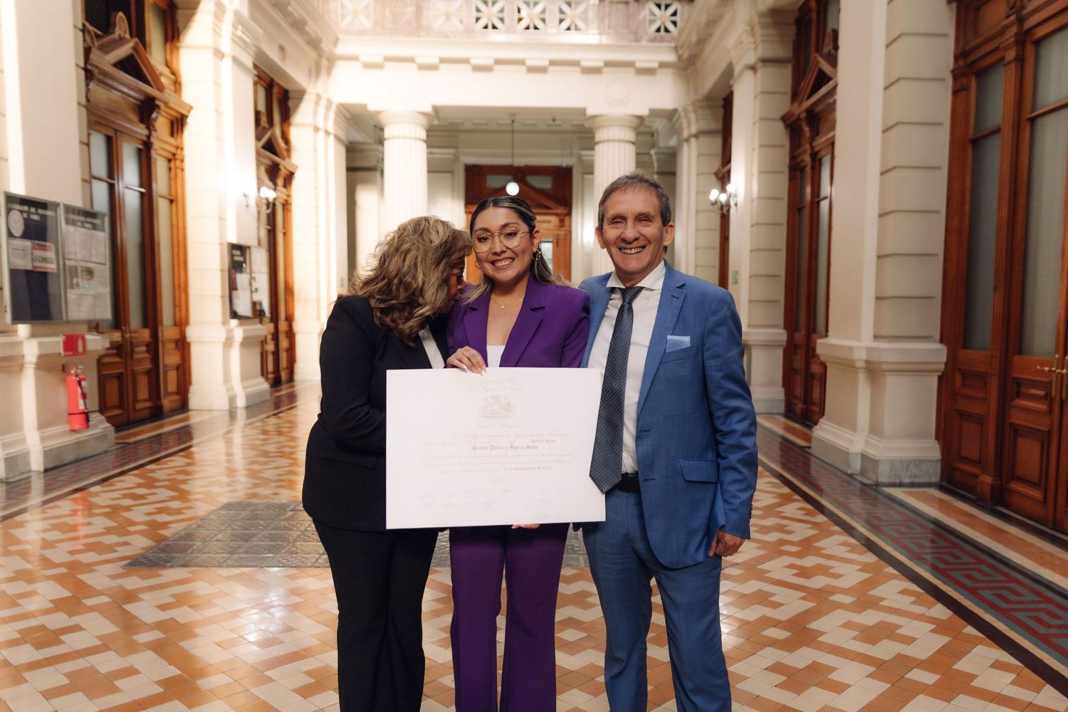 Three people posing with official certificate in ornate government building corridor.