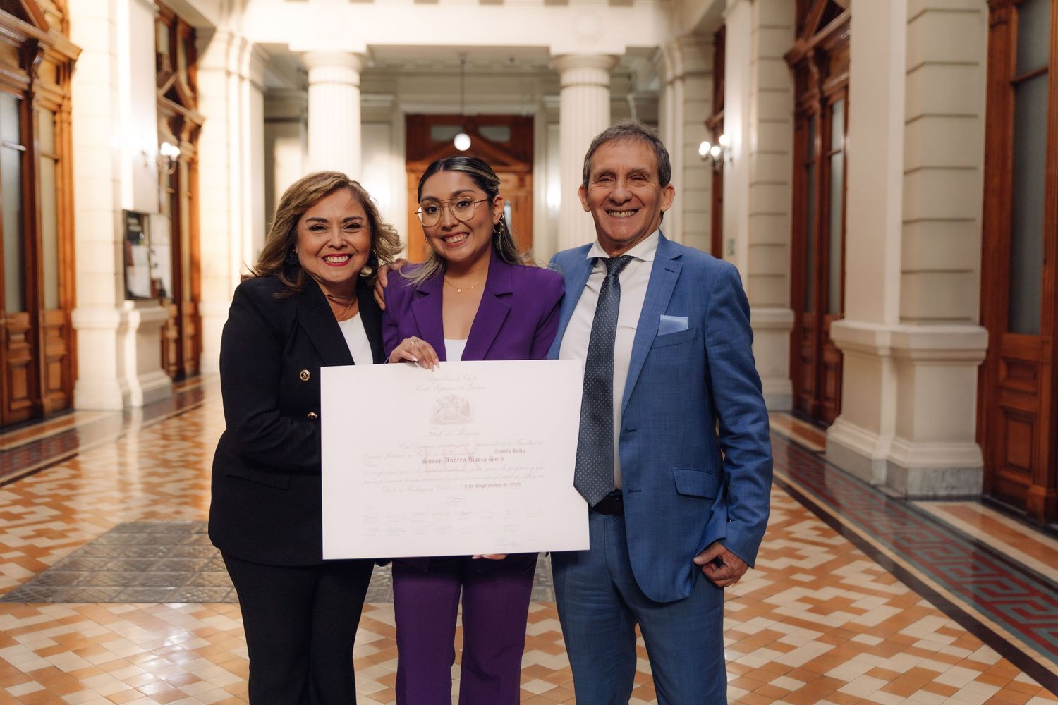 Three people posing with a large white certificate in an elegant hallway, woman in purple suit flanked by two others.