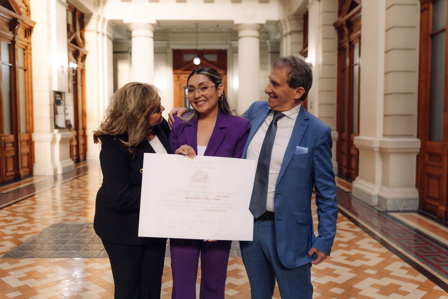 Three people smiling together holding a large white certificate in a grand building corridor with ornate architecture.