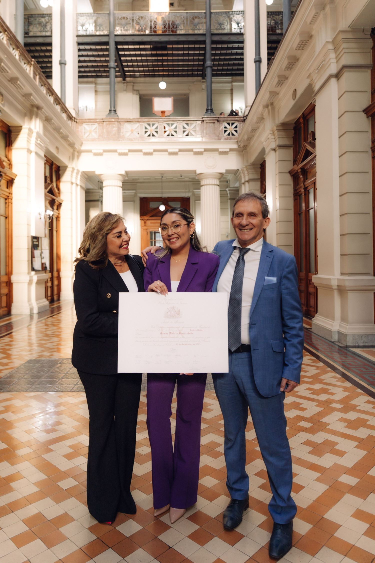Three formally dressed people proudly displaying a large white certificate in a classical building hallway.