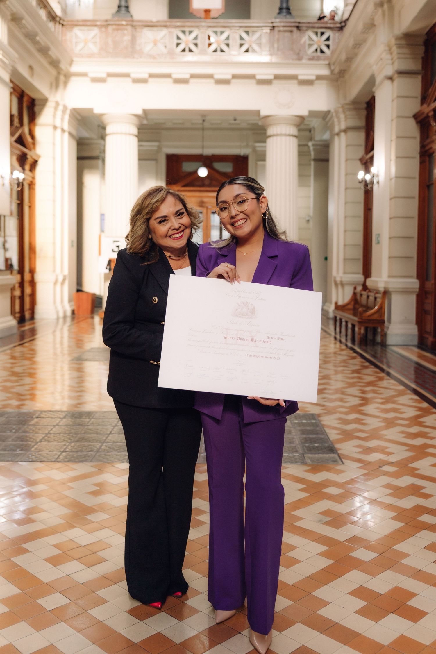 Two women in formal suits holding a large white certificate while smiling in an elegant columned hallway.