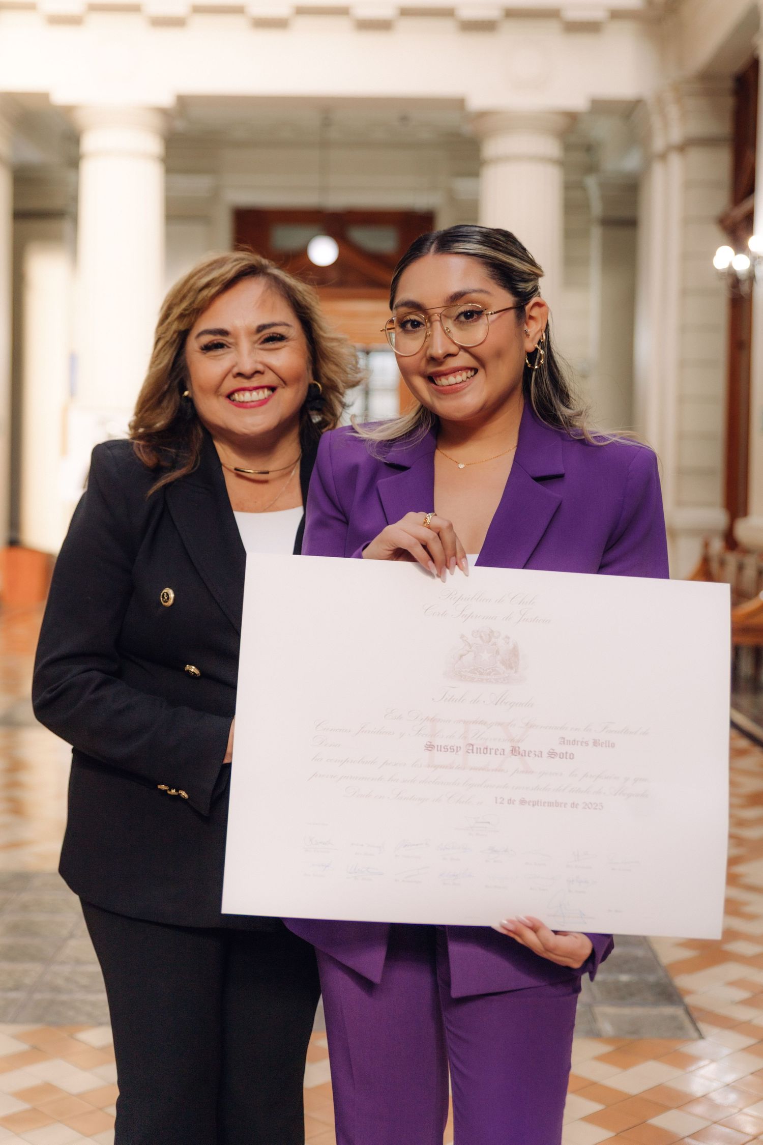 Two smiling women proudly displaying a large white certificate together in an ornate architectural hallway.