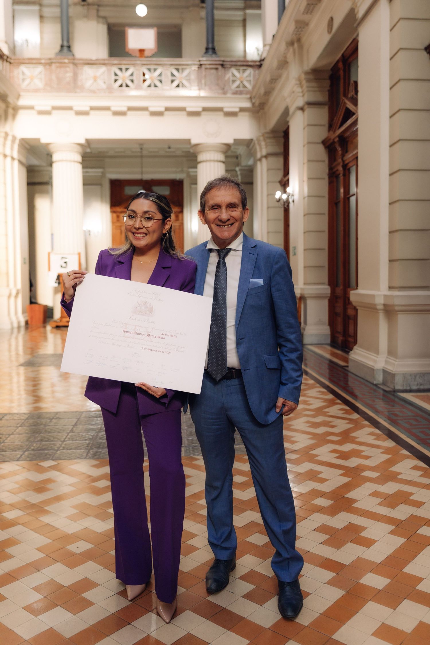 Woman in purple suit and man in blue suit posing with a large white certificate in a grand hallway.