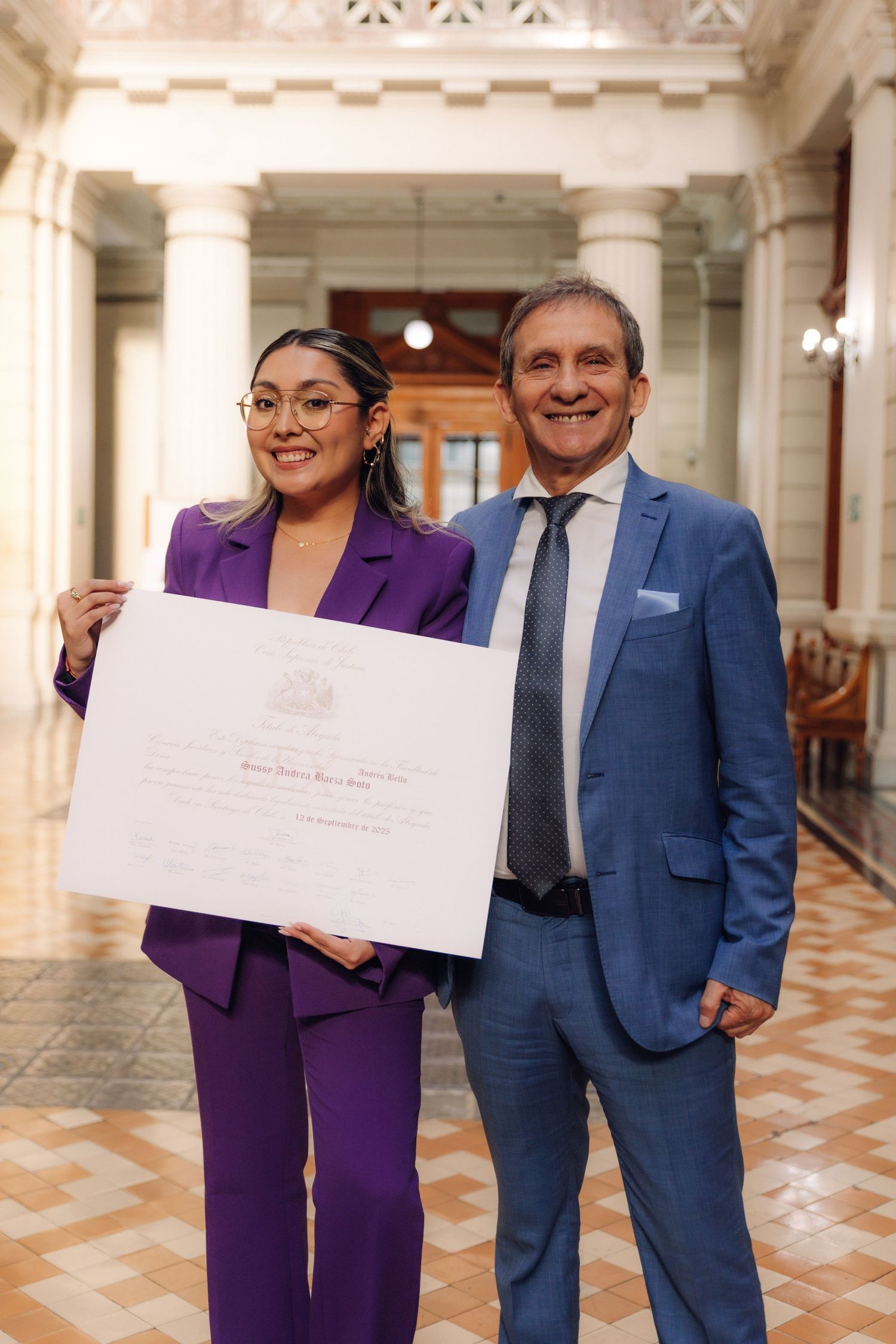 Woman in purple suit holding a large white certificate standing with a man in blue suit in elegant hall.