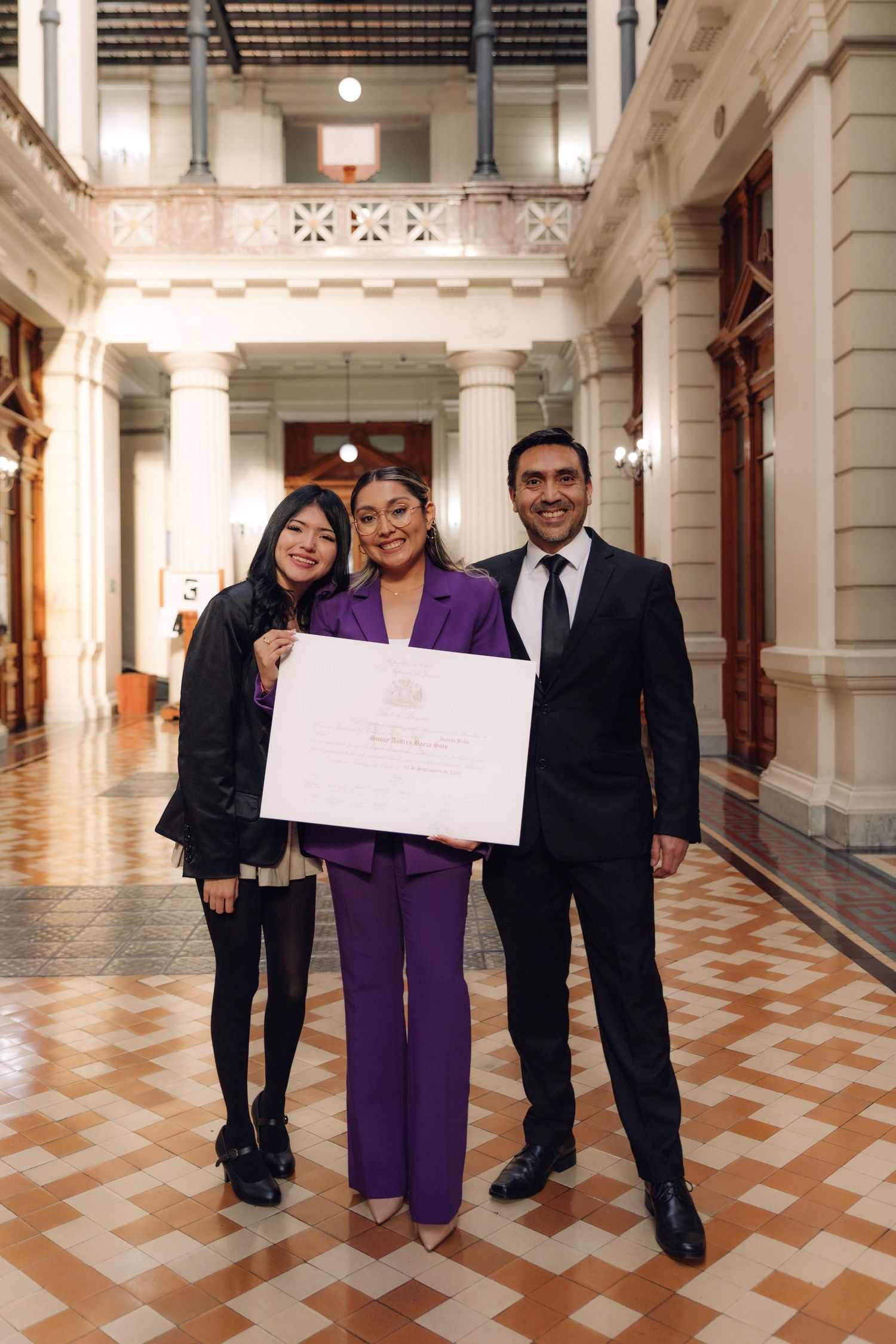 Three people smiling and holding a large white certificate in a beautifully decorated classical building hallway.